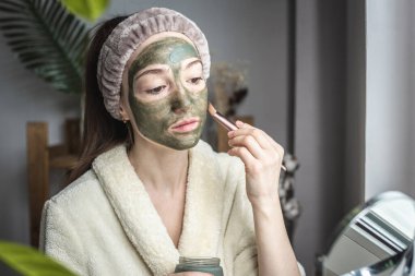 Bornozlu genç ve güzel bir kadın yüzüne yeşil bir kozmetik maske takıyor. Cilt bakımı ve yaşam tarzı kavramı.