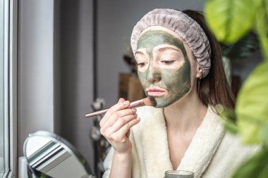 Bornozlu genç ve güzel bir kadın yüzüne yeşil bir kozmetik maske takıyor. Cilt bakımı ve yaşam tarzı kavramı.