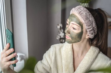 Yüzünde yeşil bir kozmetik maske olan bornozlu genç ve güzel bir kadın cep telefonunda selfie çekiyor..