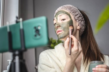 Şirin kadın blogcu, cep telefonu kamerasının önünde yüzüne kozmetik yeşil bir maske takıyor..