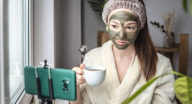 Bornozlu güzel blogcu kadın, elinde bir fincan çay ve yüzünde yeşil bir kozmetik maskeyle cep telefonu kamerasının önünde oturuyor..