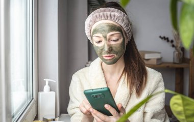 Bornozlu ve yüzünde yeşil bir kozmetik maske olan güzel bir kadın cep telefonunu kullanıyor..