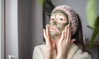 Bornozlu ve yüzünde yeşil bir maske olan güzel bir kadının portresi. Kişisel bakım, güzellik tedavileri ve doğal kozmetik kullanımı kavramı.