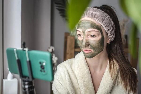 Genç, güzel bayan blogcu yüzüne bir cep telefonu kamerası önünde kozmetik yeşil maske takıyor..