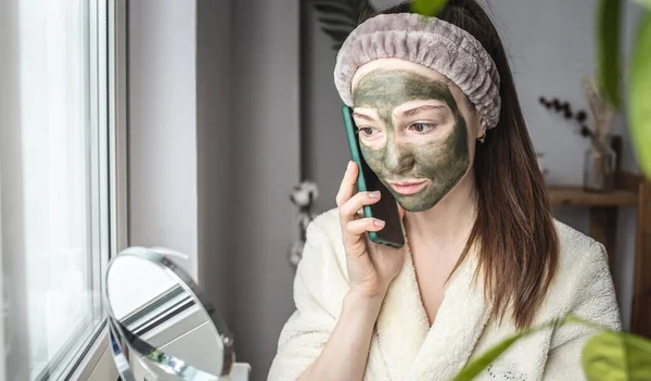 Fiatal Gyönyörű Fürdőköpenyben Zöld Kozmetikai Maszkkal Arcán Használja Mobiltelefonját — Stock Fotó