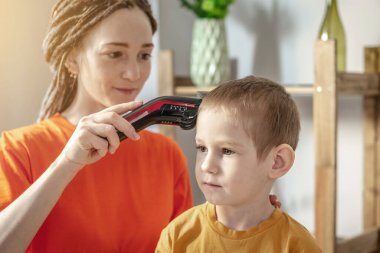 Parlak elbiseli genç modern bir anne evinde saç makasıyla küçük oğlunun saçını kesiyor. Aile ve bakım kavramı.