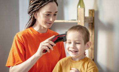 Annem evde saç makasıyla çocuğun saçını kesiyor. Küçük çocuk gülümsüyor ve keyfi yerinde. Aile ve bakım kavramı.