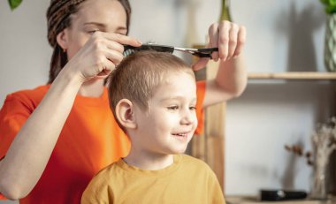 Parlak elbiseli genç bir kadın küçük bir çocuğun saçını makasla kesiyor. Aile kavramı, bakım, yaşam tarzı.