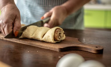Mutfakta önlüklü bir kadın lezzetli ev yapımı tarçınlı rulo yapmak için taze çiğ hamuru bıçakla nazikçe kesiyor. Pişirme süreci kavramı.