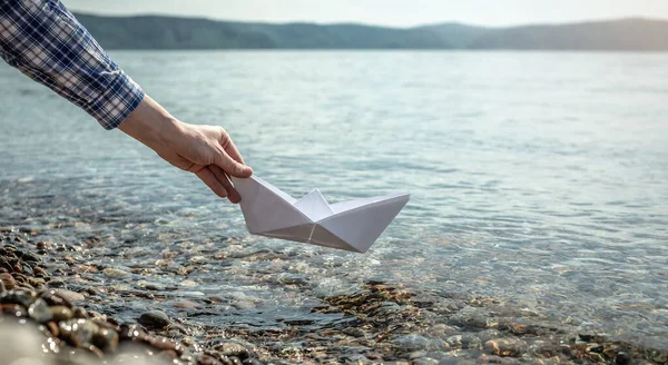 Woman\'s hand is launching a white paper boat into the clear water of a Lake Baikal with a stony bottom.