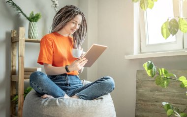 Genç modern kadın evinde bir sandalyede oturuyor ve haberleri okumak ya da diğer bilgileri görmek için bir tablet kullanıyor. Eğlence, rahatlama ve yaşam tarzı kavramı.