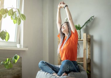 Genç modern kadın pencerenin yanındaki koltukta oturuyor ve esniyor. Sabah konsepti, sakinlik, ruh hali.