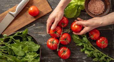 Kadının elleri olgun kırmızı bir domatesi daldan koparıyor. Siyah masada domates, roka, yeşil salata ve hafif sebze salatası pişirmek için kesim tahtası var. Üst görünüm.