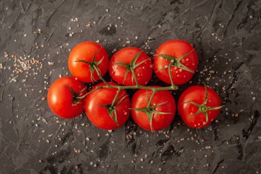Bir dalda tam olgunlaşmış kırmızı domatesler, dilimlenmiş bir domates, pembe tuz ve siyah arka planda bir kesim tahtası. Üst görünüm.