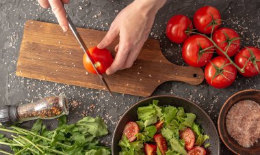 Kadınların elleri domates ve roka ile hafif sağlıklı sebze salatası pişirmek için olgun bir domates kesiyor. Siyah arkaplan ve üst görünüm.
