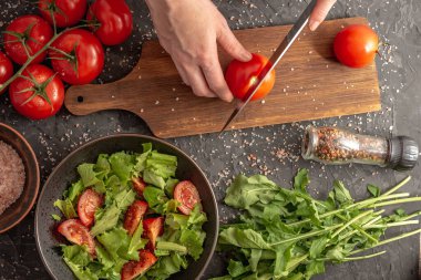 Kadınların elleri domates ve roka ile hafif sağlıklı sebze salatası pişirmek için olgun bir domates kesiyor. Siyah arkaplan ve üst görünüm.