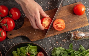 Kadınların elleri domates ve roka ile hafif sağlıklı sebze salatası pişirmek için olgun bir domates kesiyor. Siyah arkaplan ve üst görünüm.