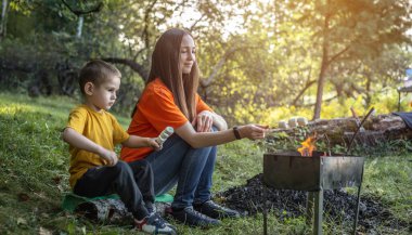 Genç anne ve küçük oğlu ateşte marşmelov kızartıyorlar. Sıcak bir sonbahar ormanında piknik, doğada bir aile eğlencesi..