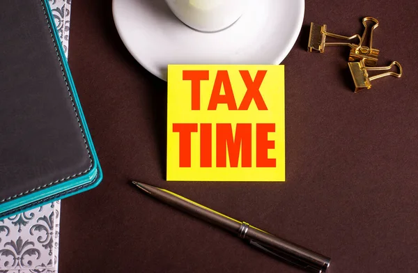 TAX TIME written on yellow paper on a brown background near a coffee cup and diaries