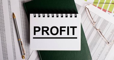 Masaüstünde raporlar, gözlükler, bir kalem, yeşil bir günlük ve içinde PROFIT yazan beyaz bir defter var. İş yeri, yakın çekim. İş konsepti