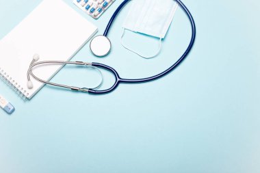 Stetoskop, boş bir not defteri ve açık mavi bir arka planda duran bir maske. Düz yatıyordu. Uzayı kopyala Tıbbi konsept