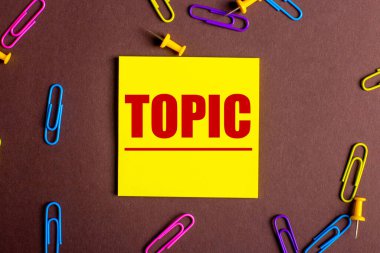 TOPIC, kahverengi arkaplanda, çok renkli ataçların yanındaki sarı etikete kırmızı ile yazılmıştır..