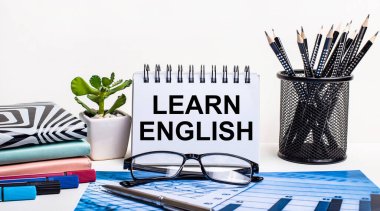 Mavi bir şema ve beyaz bir duvar, bir stantta siyah kalemler, bir çiçek, günlükler ve LeARN ENGLISH yazılı bir defter