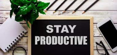 STAY PRODUCTIVE bir telefon, not defteri, gözlükler, kalemler ve yeşil bir bitkinin yanındaki siyah tahtaya beyazla yazılmıştır..
