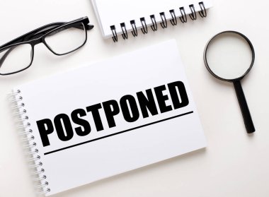 POSTPONED kelimesi defter, siyah çerçeveli gözlük ve büyüteç üzerine beyaz bir deftere yazılmıştır.