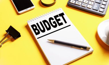 Sarı arka planda, içinde metin bütçe, kahve, hesap makinesi, telefon ve kağıtlar için bir klip olan bir defter var. İş konsepti