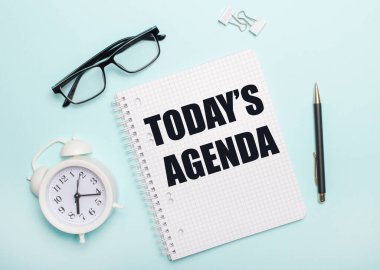 Açık mavi bir arka planda siyah gözlük ve bir kalem, beyaz bir çalar saat, beyaz ataçlar ve üzerinde TODAYS AGENDA yazan bir defter uzanıyor. İş konsepti