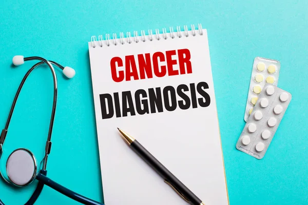 CANCER DIAGNOSIS written in a white notepad near a stethoscope, pens and pills on a light blue background. Medical concept