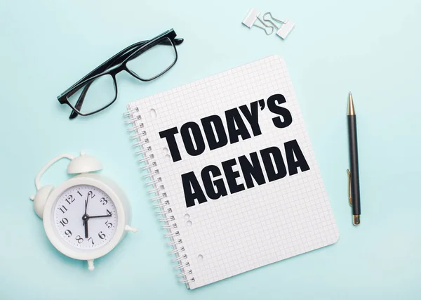 Açık mavi bir arka planda siyah gözlük ve bir kalem, beyaz bir çalar saat, beyaz ataçlar ve üzerinde TODAYS AGENDA yazan bir defter uzanıyor. İş konsepti