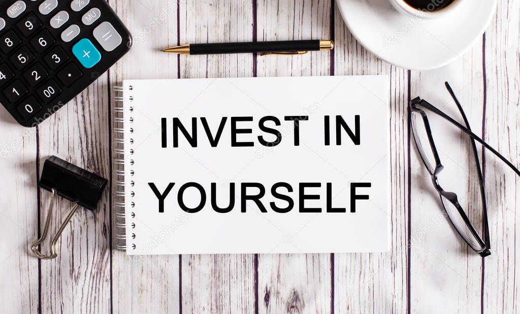 The word INVEST IN YOURSELF is written in a white notepad near a calculator, coffee, glasses and a pen. Business concept