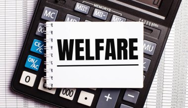 Masanın üzerinde raporlar, bir hesap makinesi ve üzerinde WELFARE yazan bir kart var. İş konsepti