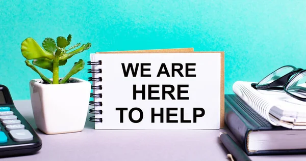 WE ARE HERE TO HELP is written on a white card next to a potted flower, diaries and calculator. Organizational concept