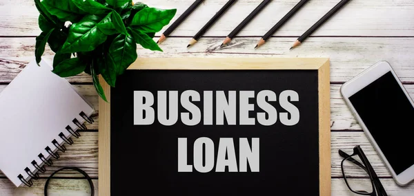 BUSINESS LOAN is written in white on a black board next to a phone, notepad, glasses, pencils and a green plant.