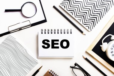 Bir ışık masasında defterler, bir büyüteç, bir çalar saat, bir gözlük ve bir kalem vardır. Ortasında da SEO yazılı bir defter var.