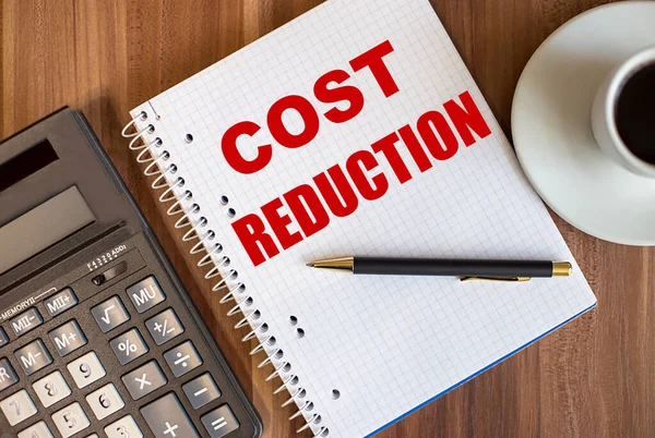 COST REDUCTION written in a white notepad near a calculator and a cup of coffee on a dark wooden background