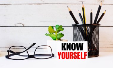 Beyaz bir arka plan gözlüğü, bir büyüteç, kalemler, bir akıllı telefon ve kendini tanı yazılı bir defter.