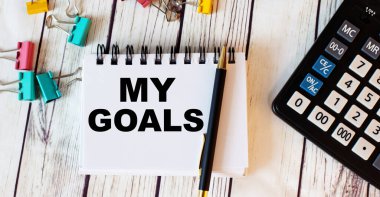 Açık ahşap bir masada hesap makinesi, çok renkli ataçlar ve kalemli bir defter ve 