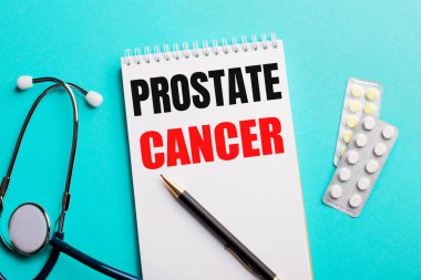 Prostate CanCER steteskopun yanında beyaz bir not defterine yazıldı. Açık mavi arka planda kalemler ve haplar. Tıbbi konsept