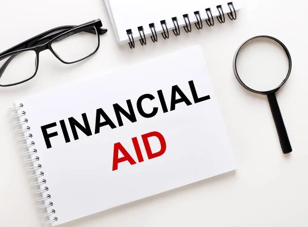 FINANCIAL AID is written in a white notebook on a light background near the notebook, black-framed glasses and a magnifying glass.