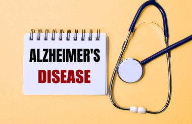 Bej bir arka plan, steteskop ve üzerinde ALZHEIMERS hastalığı yazan beyaz bir not defteri. Tıbbi konsept