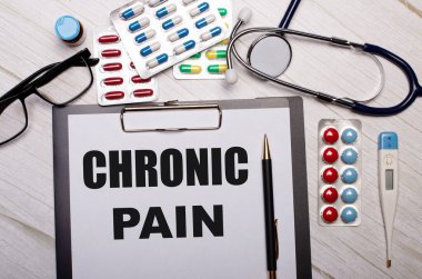 Hafif ahşap arka planda, üzerinde CHRONIC PaIN yazan kağıt, steteskop, renkli haplar, gözlük ve kalem var. Tıbbi konsept