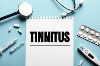 TINNITUS kelimesi steteskop, şırınga, elektronik termometre ve hapların yanındaki mavi arka planda beyaz bir not defterine yazıldı. Tıbbi konsept