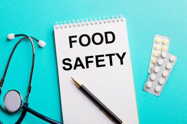 Gıda güvenliği, steteskopun yanında beyaz bir not defterine açık mavi arka planda kalemler ve haplar yazıyor. Tıbbi konsept