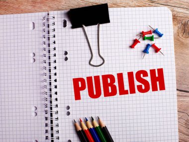 PUBLISH kelimesi çok renkli kalemler ve ahşap bir arka plandaki düğmeler yakınındaki bir deftere yazılmıştır..