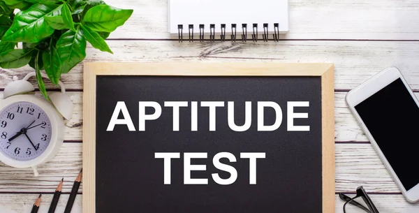 APTITUDE TEST written on a black background near pencils, a smartphone, a white notepad and a green plant in a pot