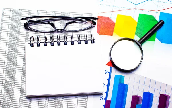 Okulary, lupa, kolorowe wykresy i biały notatnik na pulpicie. Koncepcja biznesu. Widok z góry z miejsca kopiowania — Zdjęcie stockowe